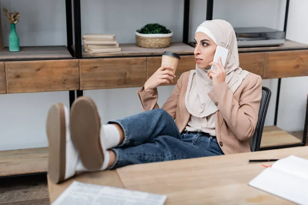 Мусульманская женщина разговаривает по смартфону, сидя с ногами на столе дома — стоковое фото
