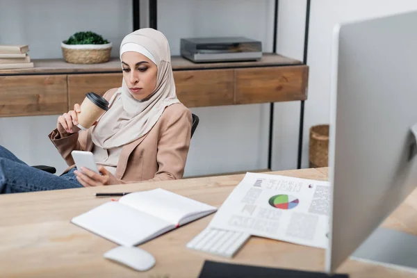 Muslimische Frau mit Coffee to go und Smartphone sitzt neben leerem Notizbuch und Tabellen auf dem Schreibtisch — Stockfoto