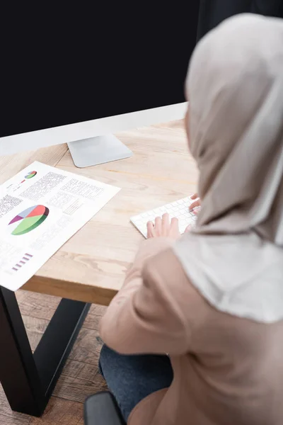 Back view of blurred muslim businesswoman typing on keyboard near infographics on desk — Stock Photo