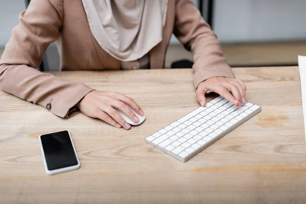 Vista recortada de la mujer usando el teclado y el ratón de la computadora mientras trabaja cerca del teléfono inteligente con pantalla en blanco - foto de stock