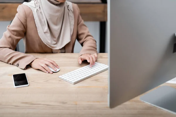 Vista cortada de mulher trabalhando no computador perto do smartphone com tela em branco na mesa — Fotografia de Stock