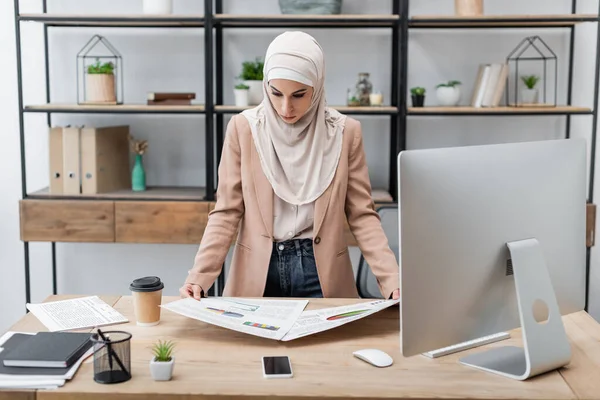 Muslim woman looking at graphs while standing near work desk at home — Stock Photo