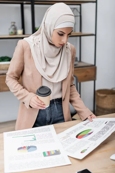 Young arabian woman looking at analytics while standing with coffee to go at work desk — Stock Photo