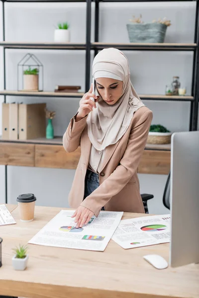 Muslimische Frau telefoniert in der Nähe von Infografiken und Computer auf dem Schreibtisch — Stockfoto