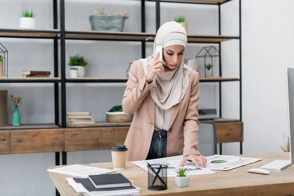 Muslim woman standing near desk with infographics and talking on mobile phone — Stock Photo
