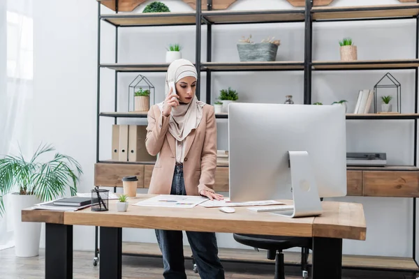 Mulher muçulmana de pé perto do computador e documentos na mesa de trabalho enquanto fala no telefone móvel — Fotografia de Stock