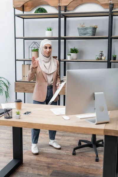 Positive muslim woman waving hand during video conference in home office — Stock Photo