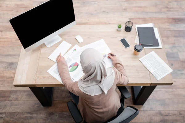 Top view of arabian woman in hijab working with charts near monitor with blank screen — Stock Photo