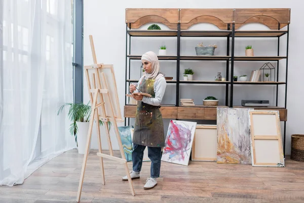 Vista completa de la mujer musulmana dibujo en caballete cerca de cuadros pintados en el estudio en casa - foto de stock