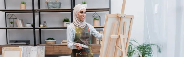 Happy woman in hijab and apron drawing on easel at home, banner — Stock Photo