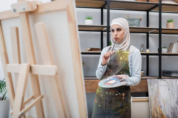 Femme arabique coûteuse avec palette et pinceau penser près de chevalet à la maison — Photo de stock