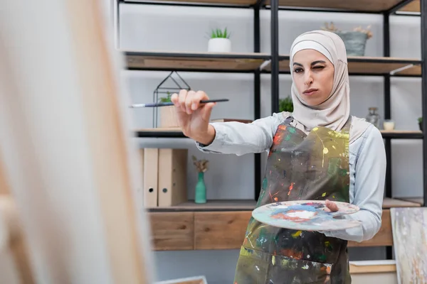 Femme musulmane avec palette et pinceau près de toile floue à la maison — Photo de stock