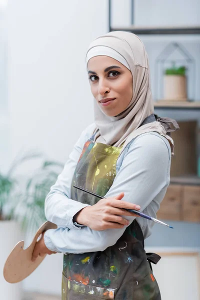 Positive muslim woman with paintbrush and palette looking at camera in home studio — Stock Photo