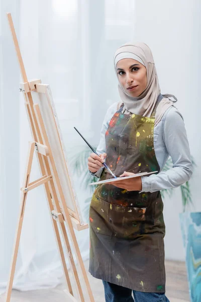 Mujer árabe con paleta y pincel mirando a la cámara cerca del caballete - foto de stock