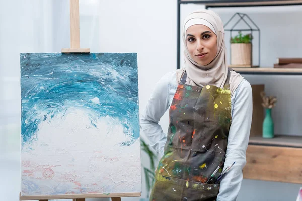 Mulher muçulmana olhando para a câmera enquanto de pé com a mão na anca perto da pintura — Fotografia de Stock