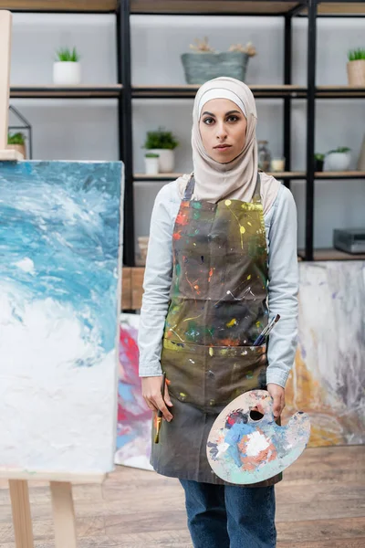 Mujer árabe con paleta mirando a la cámara cerca de la pintura en caballete - foto de stock