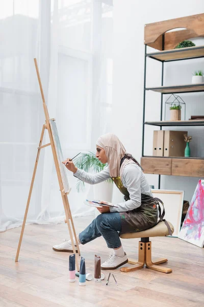 Vue latérale de la femme musulmane dessinant sur chevalet près des tubes de peinture sur le sol — Photo de stock