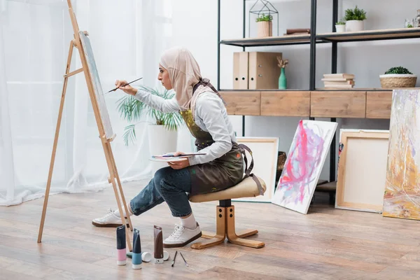 Vista laterale di donna musulmana seduta sulla sedia e disegno su cavalletto a casa — Foto stock