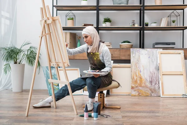 Vista completa de la mujer musulmana dibujo en caballete cerca de tubos de pintura en el suelo - foto de stock