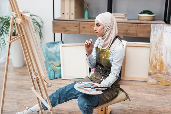 Mulher muçulmana com paleta sentado perto cavalete em estúdio em casa — Fotografia de Stock