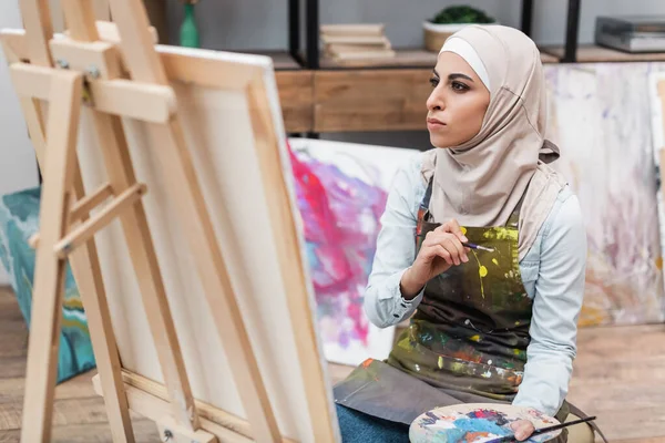 Muslim woman with palette and paintbrushes looking at canvas at home — Stock Photo