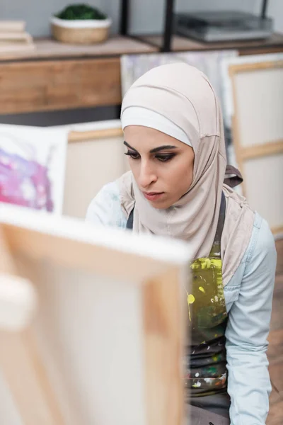 Arabian woman in hijab drawing on blurred easel at home — Stock Photo