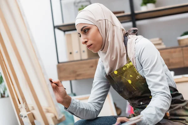 Young arabian woman in hijab painting on blurred canvas at home — Stock Photo