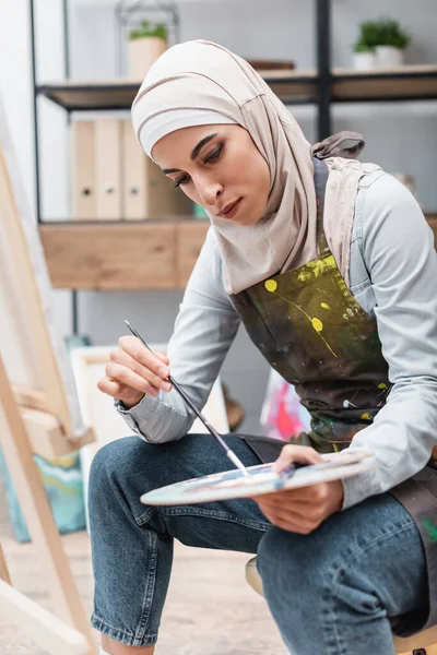 Young muslim woman with palette and paintbrush sitting near blurred easel — Stock Photo