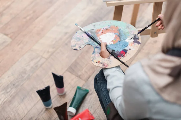 High angle view of blurred woman with palette and paintbrushes near paint tubes on floor — Stock Photo