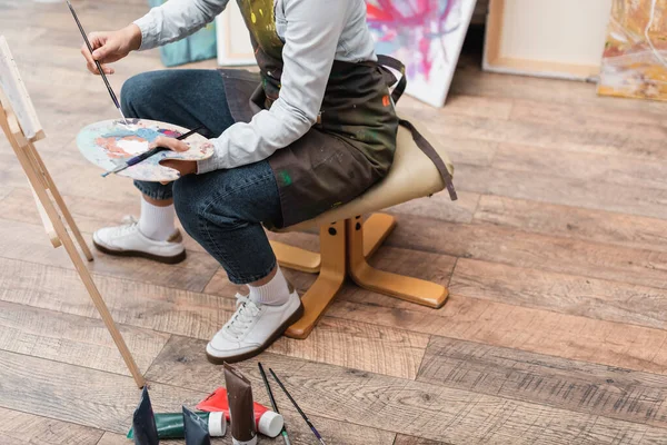 Partial view of woman with palette and paintbrushes sitting on chair near easel — Stock Photo