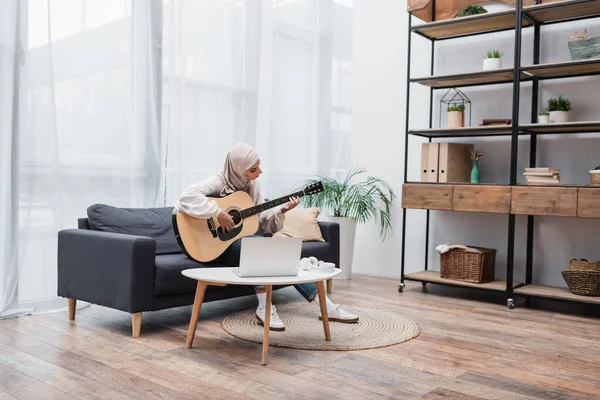 Mulher muçulmana tocando guitarra acústica perto de laptop na sala de estar moderna — Fotografia de Stock