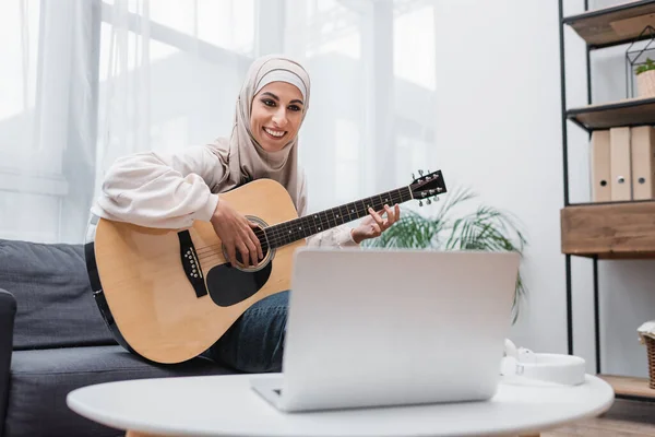 Mulher árabe alegre olhando para laptop borrado enquanto aprende a tocar guitarra em casa — Fotografia de Stock