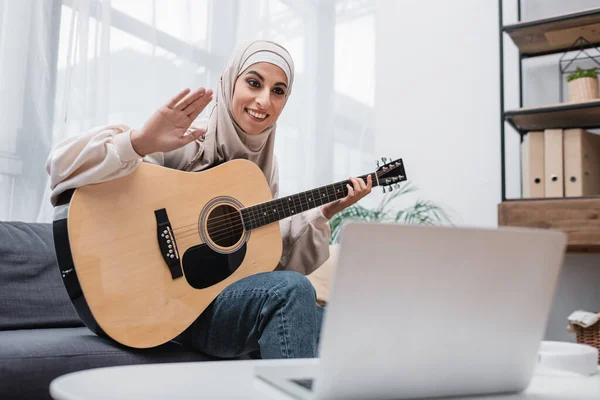 Happy muslim woman waving hand during online guitar lesson — Stock Photo