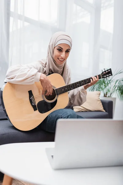 Mulher muçulmana feliz aprendendo a tocar guitarra acústica perto de laptop borrado — Fotografia de Stock