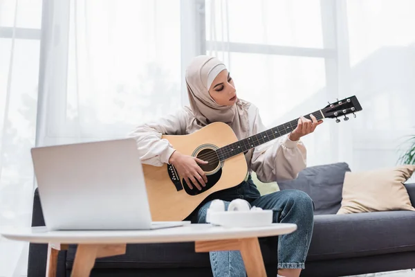 Muslim woman playing guitar while sitting on sofa during video lesson on laptop — Stock Photo