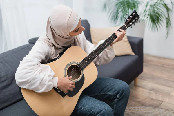 Jeune femme musulmane assise sur le canapé à la maison et jouant de la guitare — Photo de stock