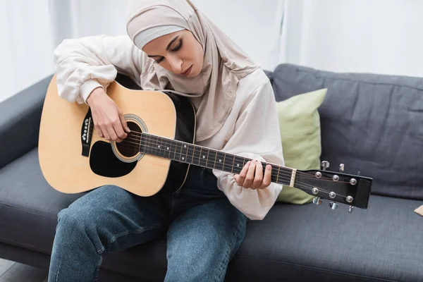 Arabo donna in hijab suonare la chitarra sul divano a casa — Foto stock