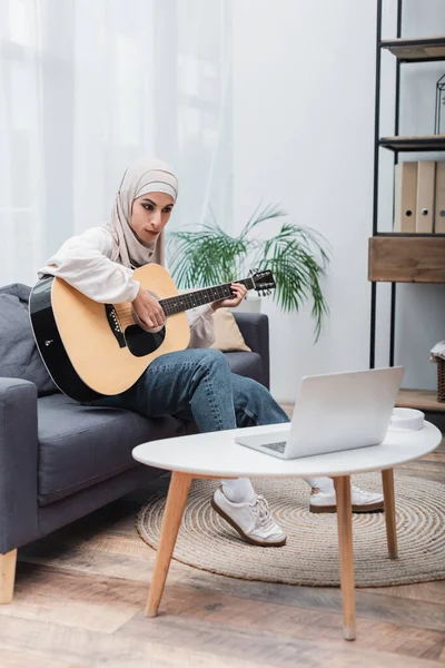 Arabian woman playing acoustic guitar in living room during online lesson on laptop — Stock Photo