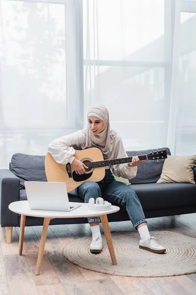 Femme musulmane souriante assise sur le canapé et apprenant à jouer de la guitare près d'un ordinateur portable — Photo de stock