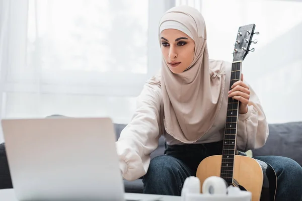 Mulher árabe no hijab segurando guitarra e olhando para laptop borrado em casa — Fotografia de Stock