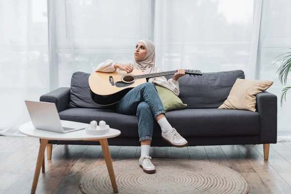 Ganzkörperansicht einer muslimischen Frau, die wegschaut, während sie auf der Couch neben dem Laptop Gitarre spielt — Stockfoto