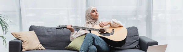 Mulher árabe olhando para longe enquanto sentado no sofá e tocando guitarra, banner — Fotografia de Stock