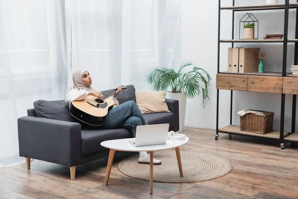 Mulher árabe no hijab tocando guitarra no sofá perto do laptop — Fotografia de Stock