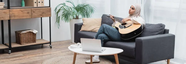 Jovem mulher muçulmana tocando guitarra enquanto sentado no sofá perto do laptop na sala de estar, banner — Fotografia de Stock