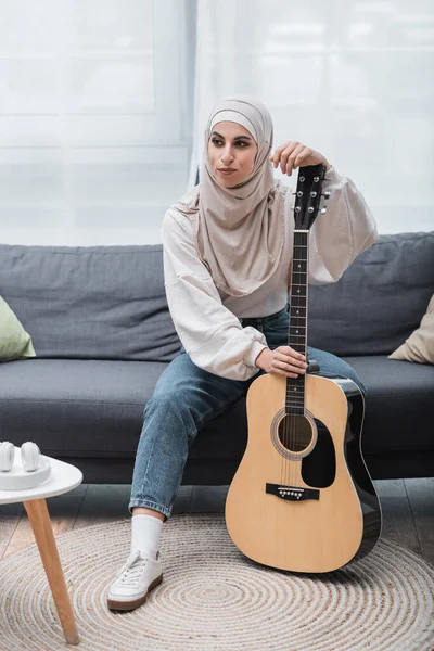 Мусульманская женщина смотрит в сторону, сидя на диване с акустической гитарой — стоковое фото