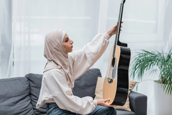 Vista lateral de la mujer musulmana mirando la guitarra mientras está sentado en el sofá en casa - foto de stock