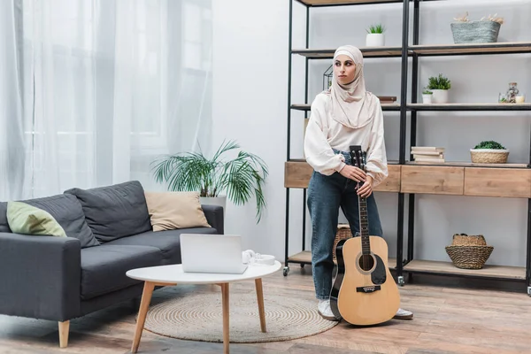 Ganzkörperansicht einer arabischen Frau, die mit Akustikgitarre im modernen Wohnzimmer steht — Stockfoto