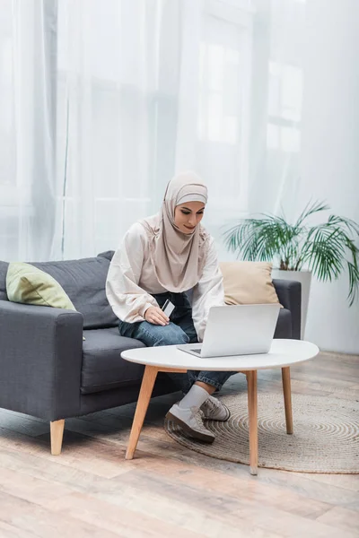 Femme musulmane assis sur le canapé avec carte de crédit près d'un ordinateur portable sur la table basse — Photo de stock
