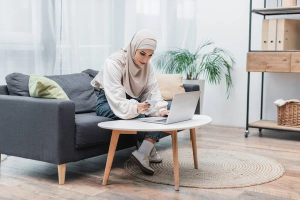 Muslim woman in hijab holding credit card and using laptop on couch at home — Stock Photo