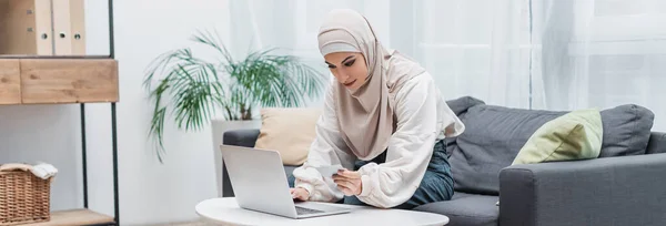 Joven mujer musulmana sosteniendo tarjeta de crédito durante las compras en línea en casa, bandera - foto de stock